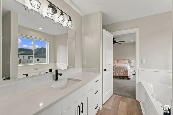 Bathroom featuring a white vanity with a light-toned countertop, undermount sink, matte black faucet, and industrial-style multi-light fixture, accompanied by wood-finish flooring and a built-in soaking tub