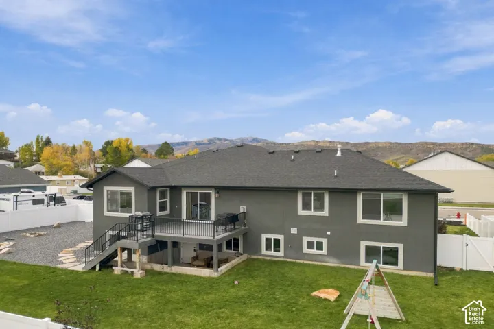Exterior featuring a gray stucco facade, elevated deck with metal railings, gravel and grass landscaping, white vinyl perimeter fencing, and a mountain backdrop