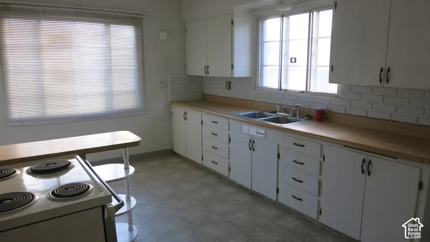 Kitchen featuring white cabinetry, light-toned countertops, a dual-basin stainless steel sink, tile backsplash, and a built-in electric cooktop
