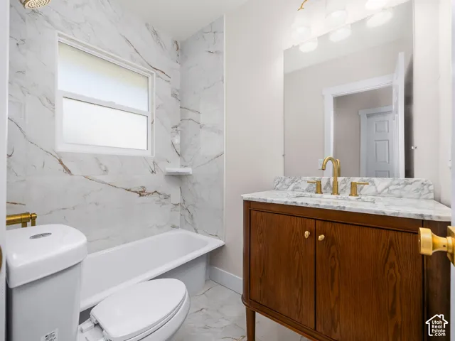 Bathroom featuring a wood-finish vanity with a stone countertop, an integrated sink, brushed gold-tone hardware, and tub surround tiled in a neutral stone pattern
