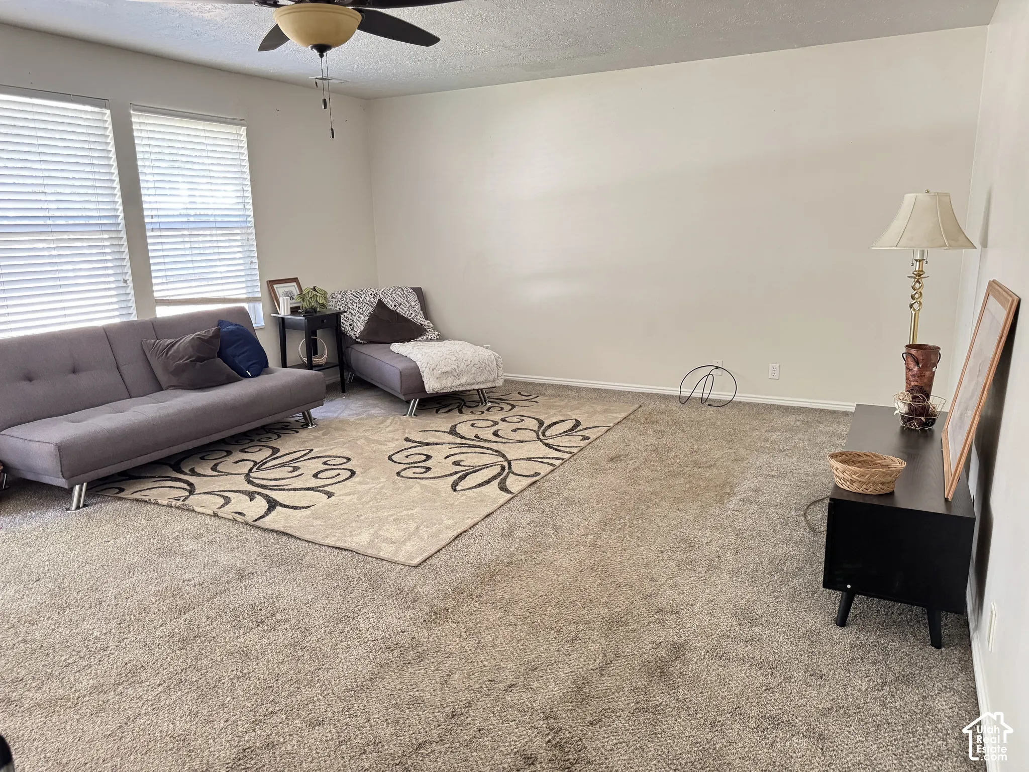 Spacious room featuring neutral carpeting, high-reaching walls, dual-pane windows with horizontal blinds, and a mounted ceiling fan with integrated lighting