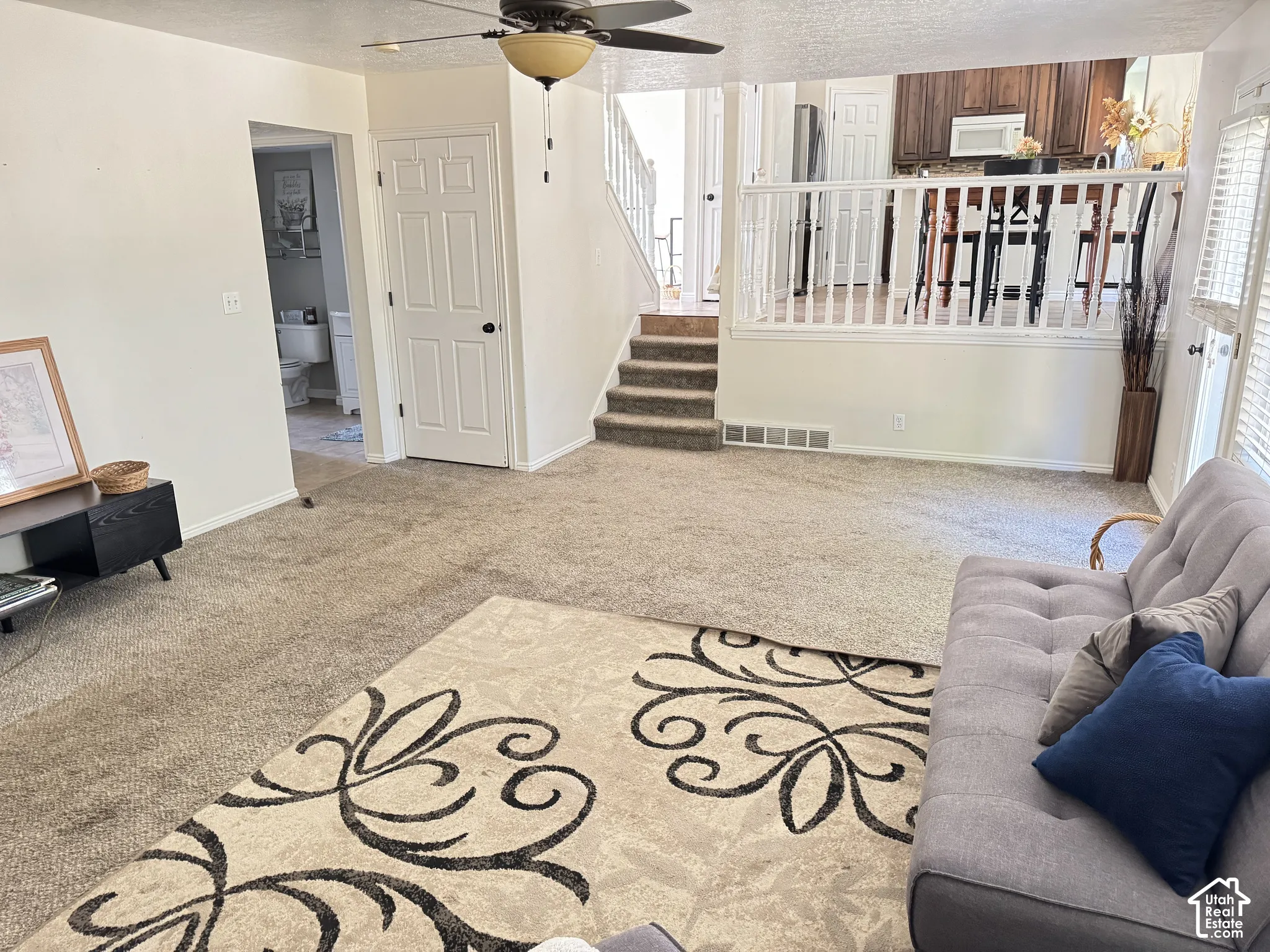 Open-concept interior featuring carpeted flooring, a ceiling-mounted light fixture with a fan, a staircase with a wooden balustrade, and a raised kitchen area with dark cabinetry
