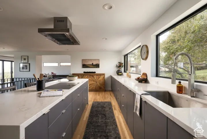 Kitchen featuring matte charcoal flat-panel cabinetry, light-toned stone-finish countertops, a central island with a flush-mount cooktop, a stainless steel industrial-style faucet, and a contemporary box-style range hood
