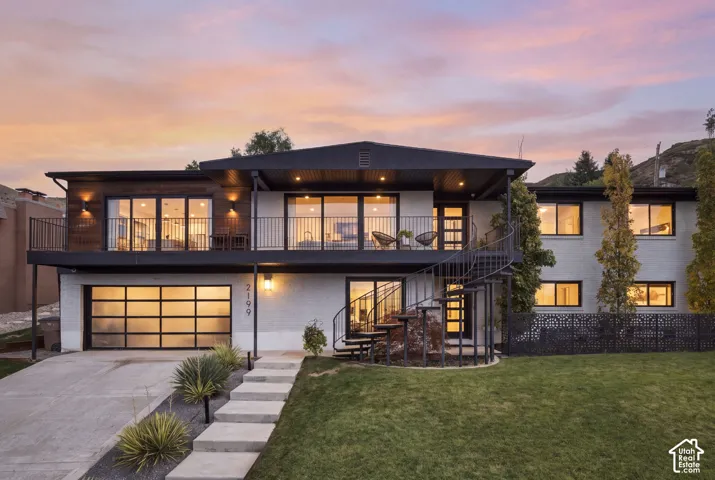 Mid-century modern architectural exterior featuring painted brick masonry, horizontal wood-finish siding, a full-width upper-level balcony with metal railing, a prominent black metal spiral staircase, and a contemporary glass-paneled garage door