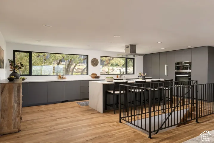 Modern kitchen featuring matte cabinetry, stone-topped central island, built-in double wall ovens, stainless steel range hood, and wood-finish flooring