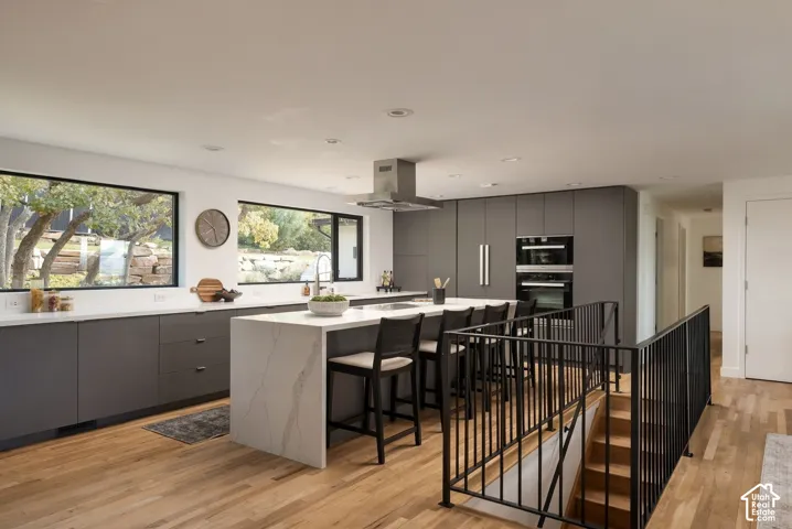 Modern kitchen featuring matte grey cabinetry, a waterfall-edge island with stone-finish countertop, stainless steel overhead range hood, built-in double wall ovens, and wood-finish flooring