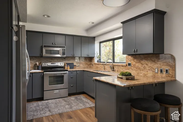 Kitchen featuring dark-toned cabinetry, stone countertops with integrated backsplash, stainless steel appliances, and wood-finish flooring