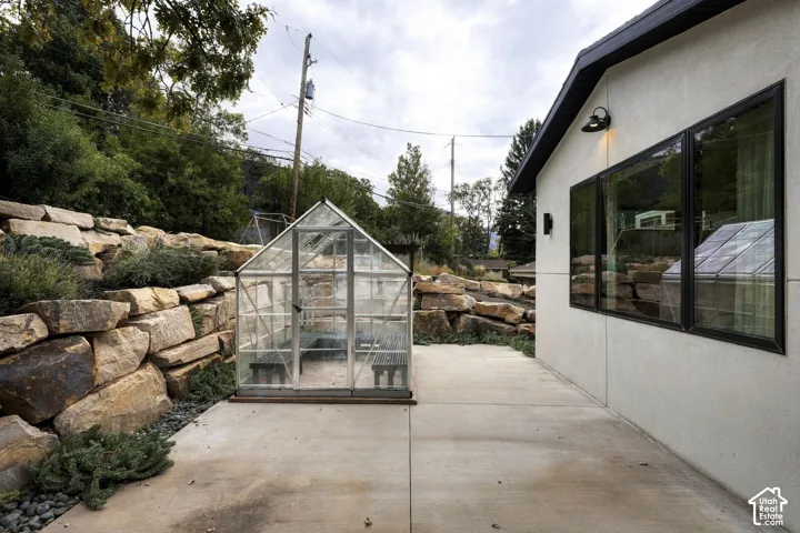 Concrete patio featuring a detached glass-paneled greenhouse, natural stone boulder retaining walls, and exterior wall-mounted lighting