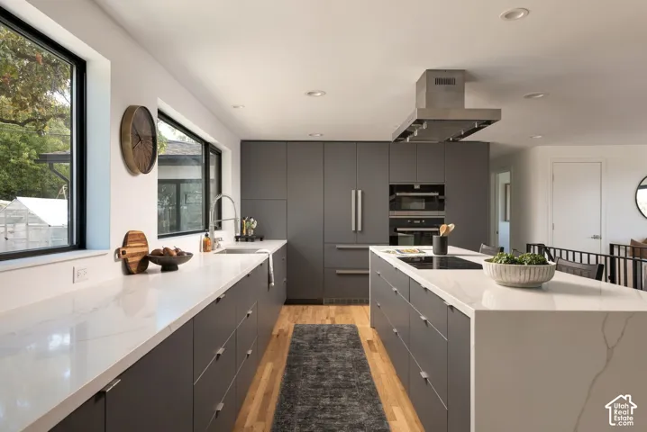 Kitchen featuring matte charcoal flat-panel cabinetry, white veined countertops, a large center island with an integrated induction cooktop, stainless steel wall-mounted range hood, and wood-finish flooring
