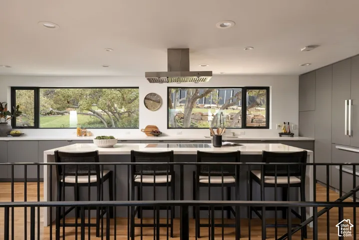 Kitchen featuring flat-panel gray cabinetry, a central island with a solid-surface countertop, an integrated cooktop, a stainless steel chimney-style range hood, and wood-finish flooring