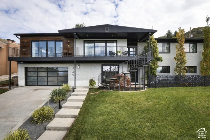Mid-century modern exterior featuring painted brick masonry, wood-finish siding, a sprawling upper-level balcony with metal railing, a prominent spiral staircase, and a multi-panel glass garage door