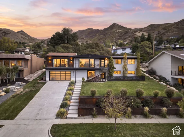 Multi-level contemporary exterior featuring a translucent multi-panel garage door, cantilevered balcony with metal railing, weathered steel retaining walls, and tiered hardscaping set against a mountain backdrop