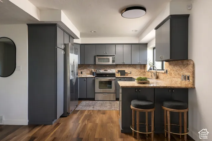Kitchen featuring dark gray cabinetry, stone-surface countertops with matching backsplash, wood-finish flooring, and stainless steel appliances