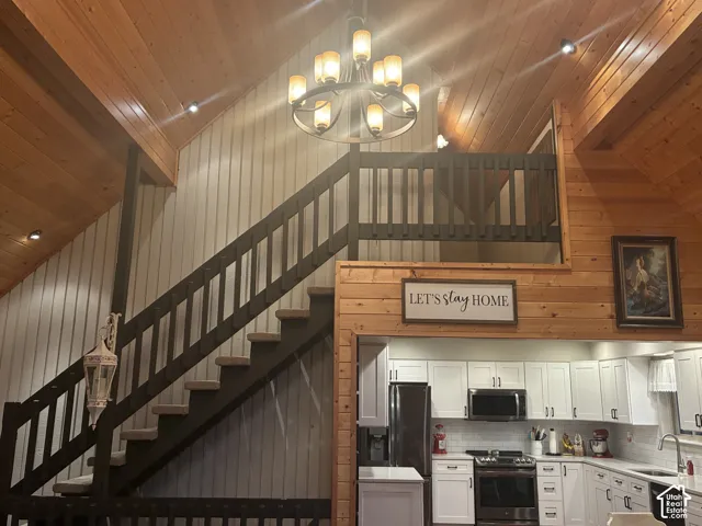 Vaulted ceilings with exposed wood beams, wood-paneled walls, a large circular chandelier, and a custom staircase featuring a dark-finish railing