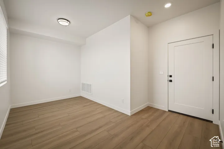Neutral-toned interior space featuring wood-finish flooring, a solid-panel door with matte black hardware, and recessed ceiling lighting