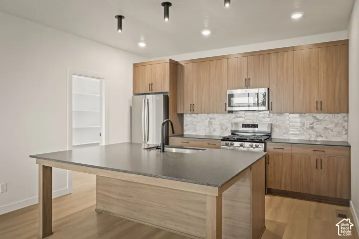 Kitchen featuring flat-panel wood-finish cabinetry, a central island with a solid-surface countertop, stone-tiled backsplash, stainless steel appliances, and matte black plumbing fixtures