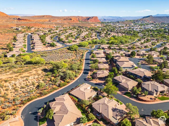 Aerial perspective of a residential community featuring paved roadways, low-profile tiled roofs, desert landscaping, and a backdrop of red rock mountainous terrain