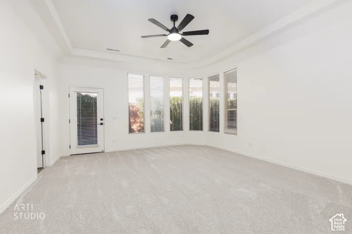 Tray ceiling, matte black ceiling fan with integrated light, neutral carpeting, and a series of tall windows with horizontal blinds