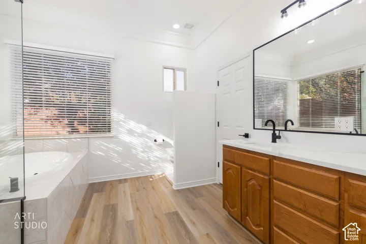 Bathroom featuring wood-finish flooring, a jetted soaking tub with tile surround, a wood vanity with a solid-surface countertop, matte black fixtures, and a large framed mirror