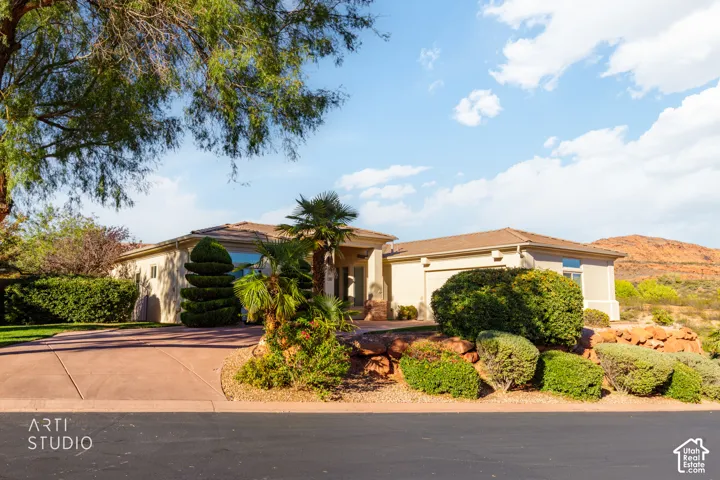 Single-level residence with stucco exterior, concrete tile roof, multi-car garage, and desert-inspired landscaping featuring mature trees and rock hardscaping