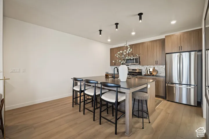 Kitchen featuring wood-finish cabinetry, a central island with a stone-top surface, stainless steel appliances, a tile backsplash, and recessed cylinder lighting