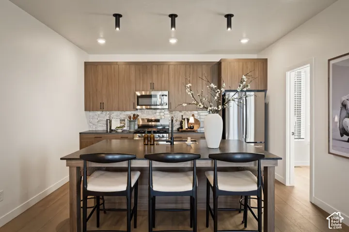Kitchen featuring flat-panel wood-finish cabinetry, stainless steel appliances, patterned tile backsplash, dark solid-surface countertop, and wood-finish flooring