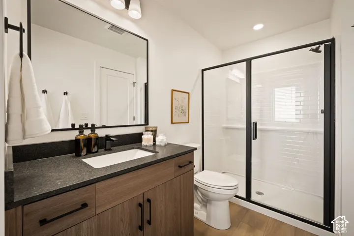 Bathroom featuring a vanity with dark stone-finish countertop, under-mount sink, matte black hardware, and wood-finish cabinetry, paired with a walk-in shower enclosed by a black-framed glass door with white subway tile surround and wood-finish flooring
