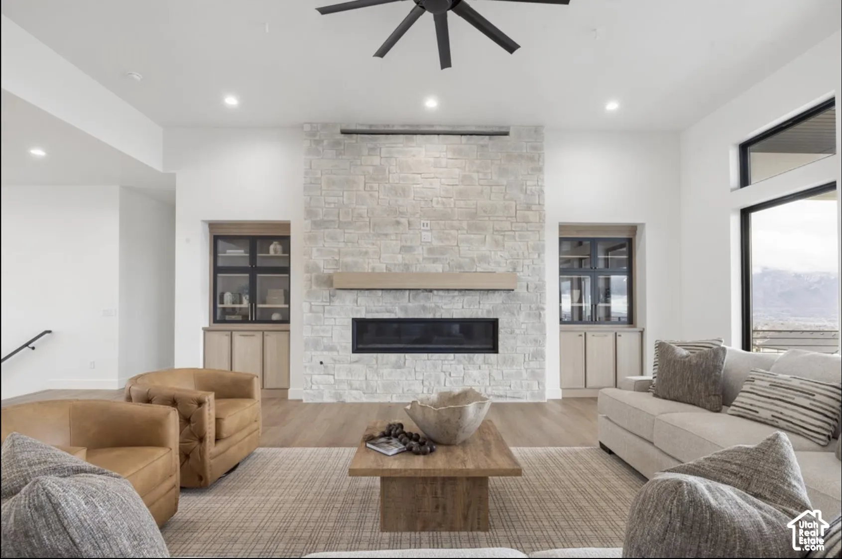 Floor-to-ceiling stone fireplace featuring a linear hearth and wood mantel, flanked by built-in cabinetry with glass-front shelving, wood-finish flooring, and a modern multi-blade ceiling fan