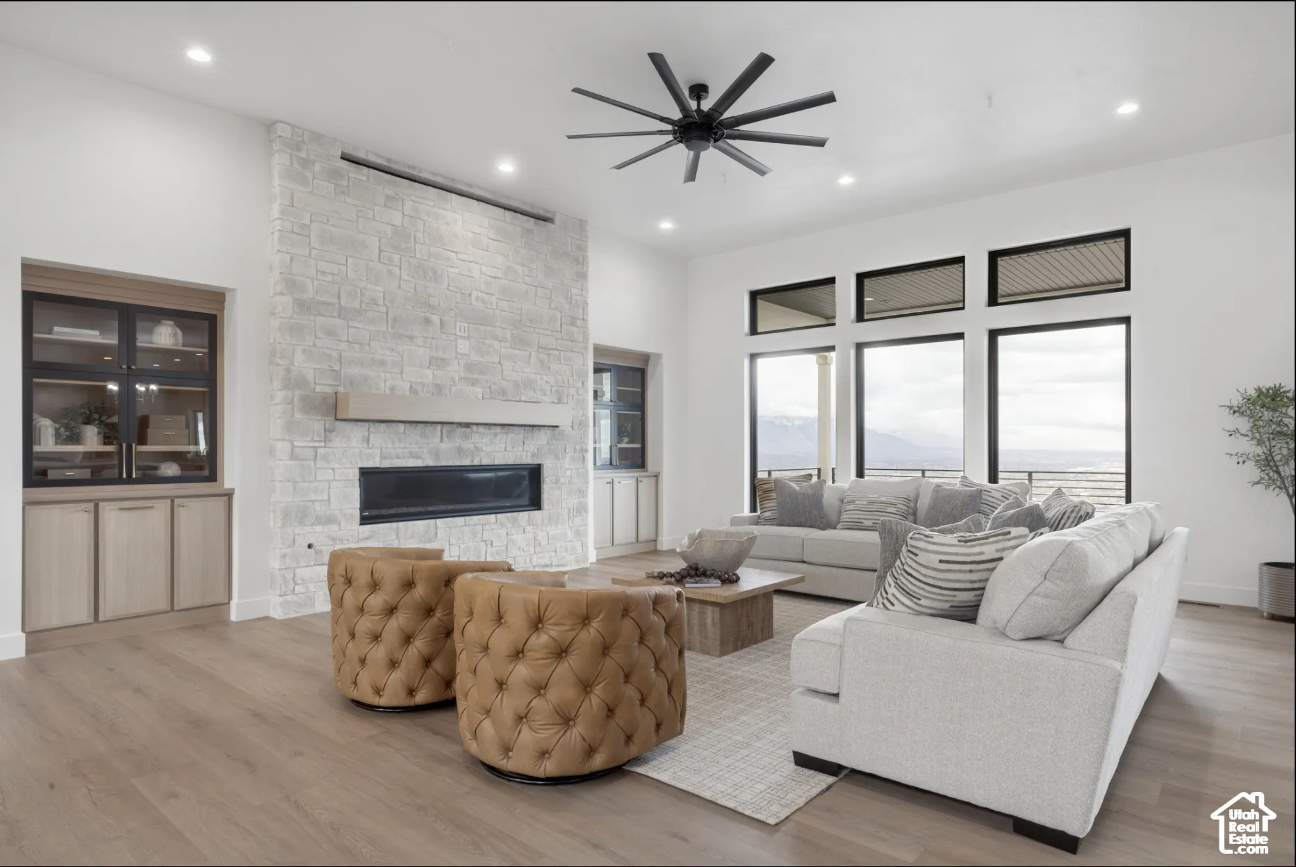 Floor-to-ceiling light-colored stone fireplace with linear hearth, wood-finish flooring, built-in cabinetry with glass-front shelving, large windows with transom accents, and multi-blade ceiling fan