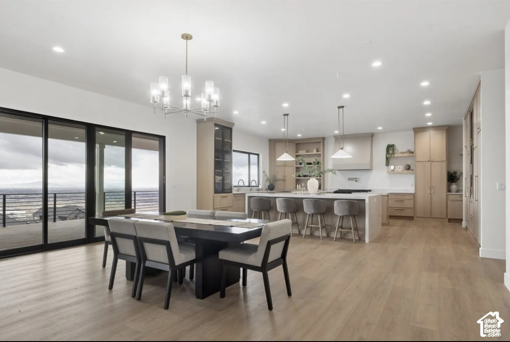 Open-concept kitchen and dining area featuring custom light wood cabinetry, a central island with waterfall-edge stone countertop, recessed lighting, chandelier and pendant fixtures, and multi-panel sliding glass doors