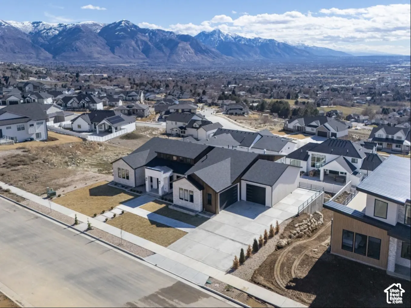 Aerial perspective of a multi-gabled residence featuring a white stucco and wood-paneled exterior, a multi-car garage, concrete driveway, and prominent mountain backdrop