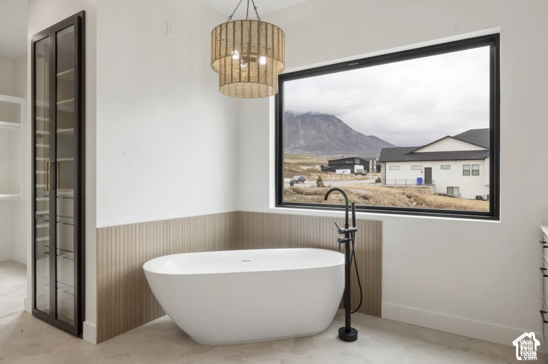 Freestanding soaking tub with matte black floor-mounted faucet, integrated vertical-slatted wall paneling, large picture window with mountain backdrop, and full-height glass-front built-in cabinetry