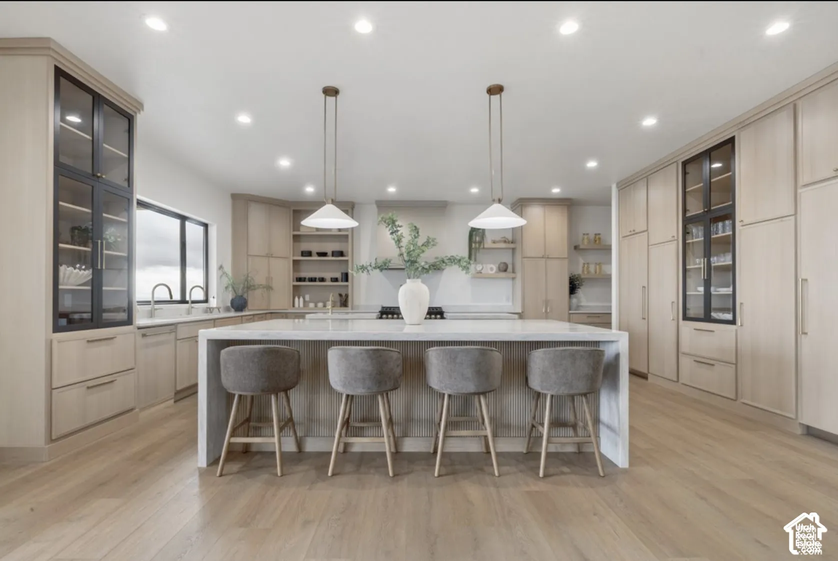 Kitchen featuring custom light-finish cabinetry, floor-to-ceiling glass-paneled display cupboards, a waterfall-edge stone island, recessed lighting, and wood-finish flooring