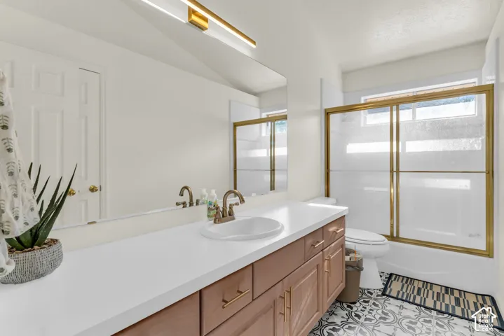 Bathroom featuring a wood-finish vanity with a solid-surface countertop, an oval under-mount sink, brass-tone hardware, and a large wall-to-wall mirror with linear vanity lighting above a bathtub-shower combination with a brass-tone sliding enclosure