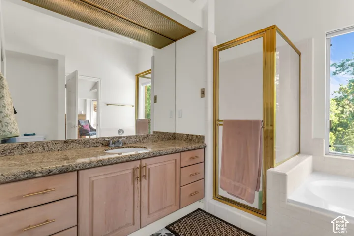 Bathroom featuring a stone-surface vanity with undermount sink, custom wood cabinetry, a glass-enclosed shower with gold-tone metal trim, and a built-in soaking tub