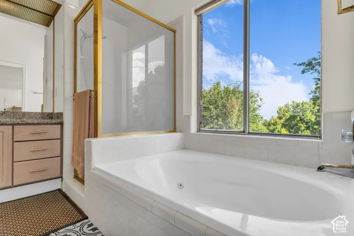 Soaking tub with tile surround, glass-enclosed shower with gold-tone frame, vanity featuring stone countertop, and large window