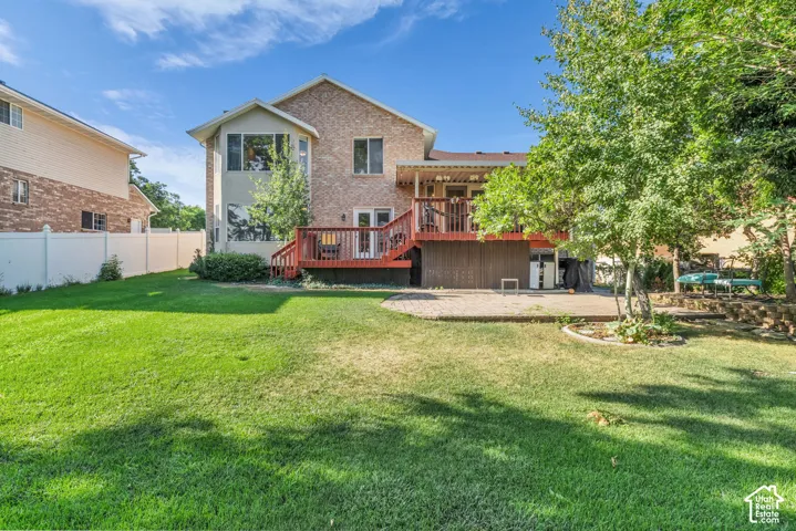 Rear exterior featuring multi-level brick facade, elevated wood deck with railings, ground-level stone paver patio, expansive lawn, and white privacy fencing