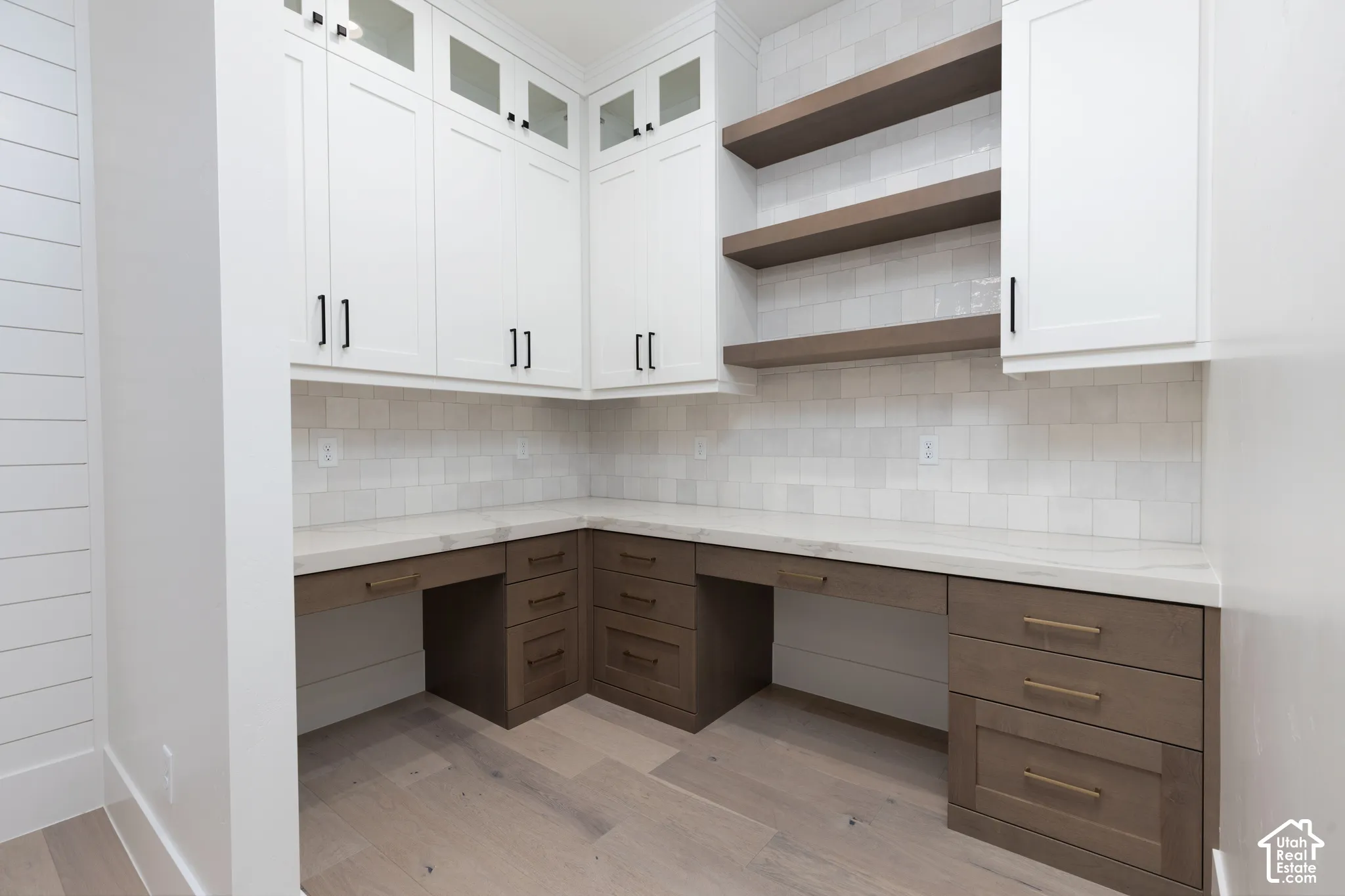 Built-in cabinetry featuring white upper cabinets with glass-paneled doors, wood-finish lower drawers, stone-surface countertops, square tile backsplash, and floating wood shelving