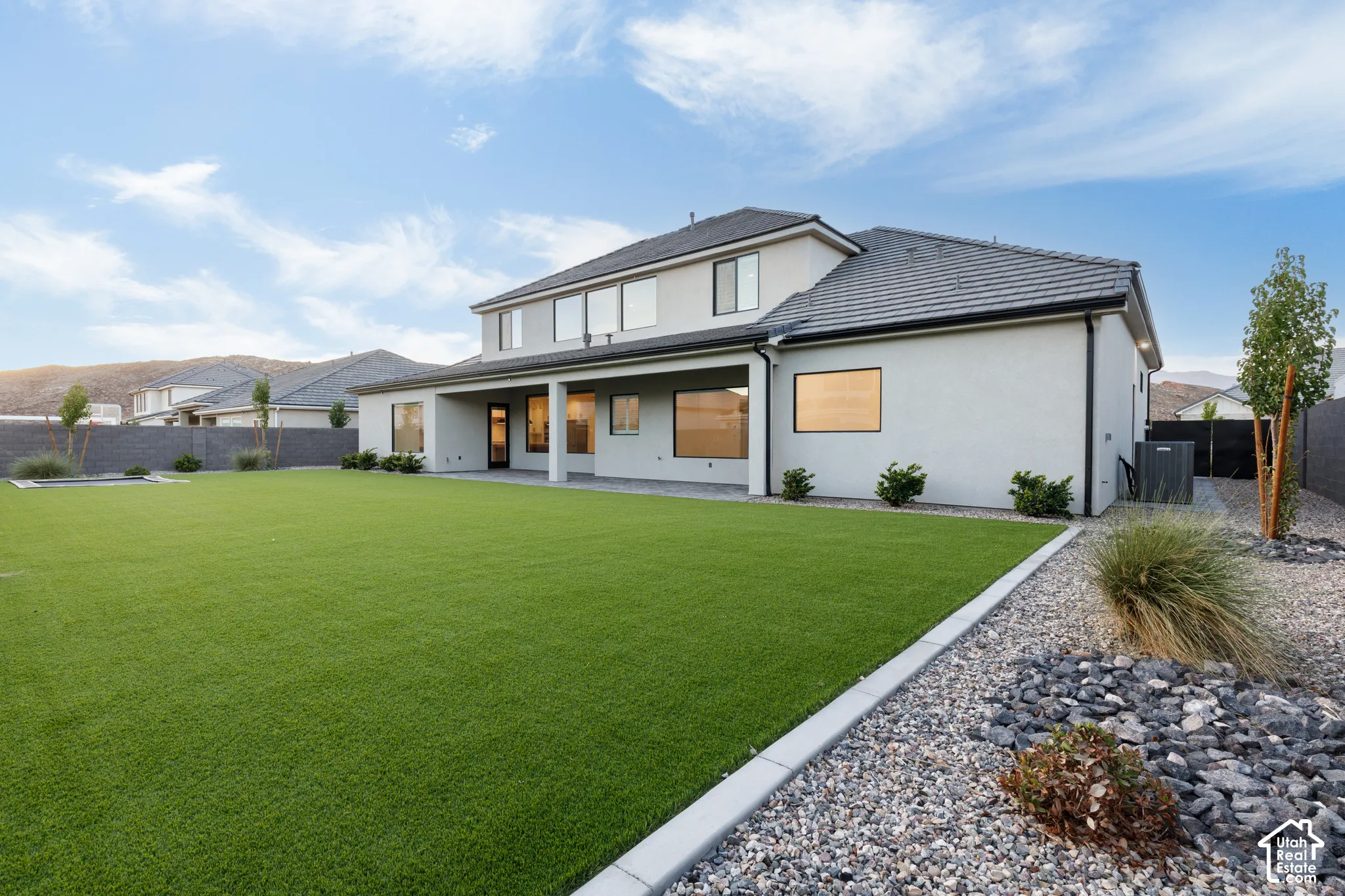 Rear exterior featuring a covered patio, synthetic turf lawn, stone-accented hardscaping, and perimeter masonry fencing