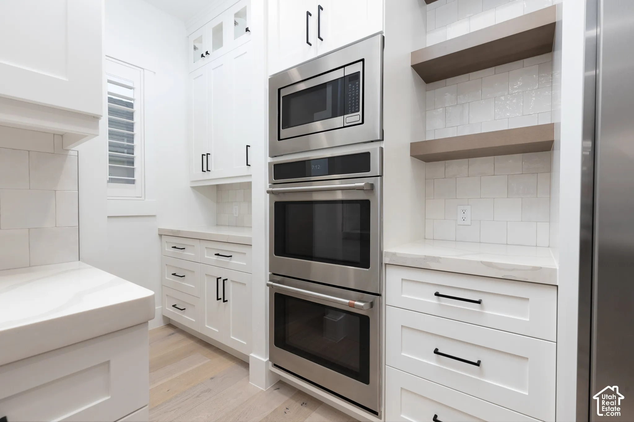 Custom white cabinetry with matte black hardware, stainless steel built-in double ovens and microwave, stone countertops, square-profile tile backsplash, and wood-finish flooring
