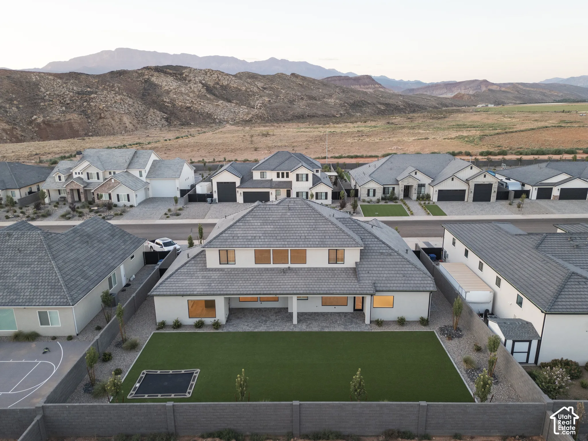 Two-story residence featuring a tile roof, covered rear patio, expansive synthetic turf lawn, stone-paved hardscaping, and perimeter masonry fencing, set against a mountain backdrop