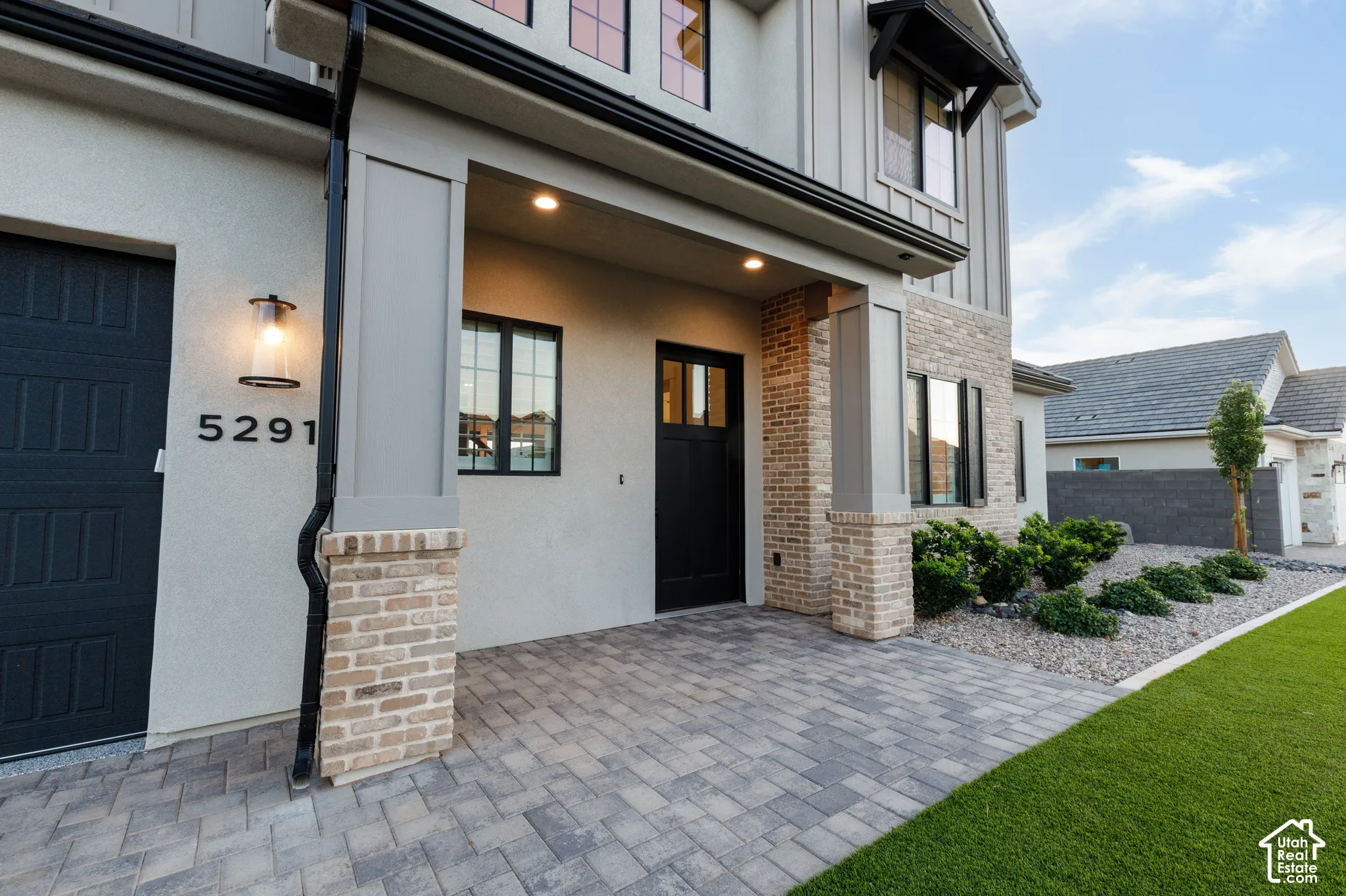 Exterior facade featuring a combination of stucco and masonry, covered entryway with paver flooring, modern panel door, and low-maintenance xeriscaped front yard