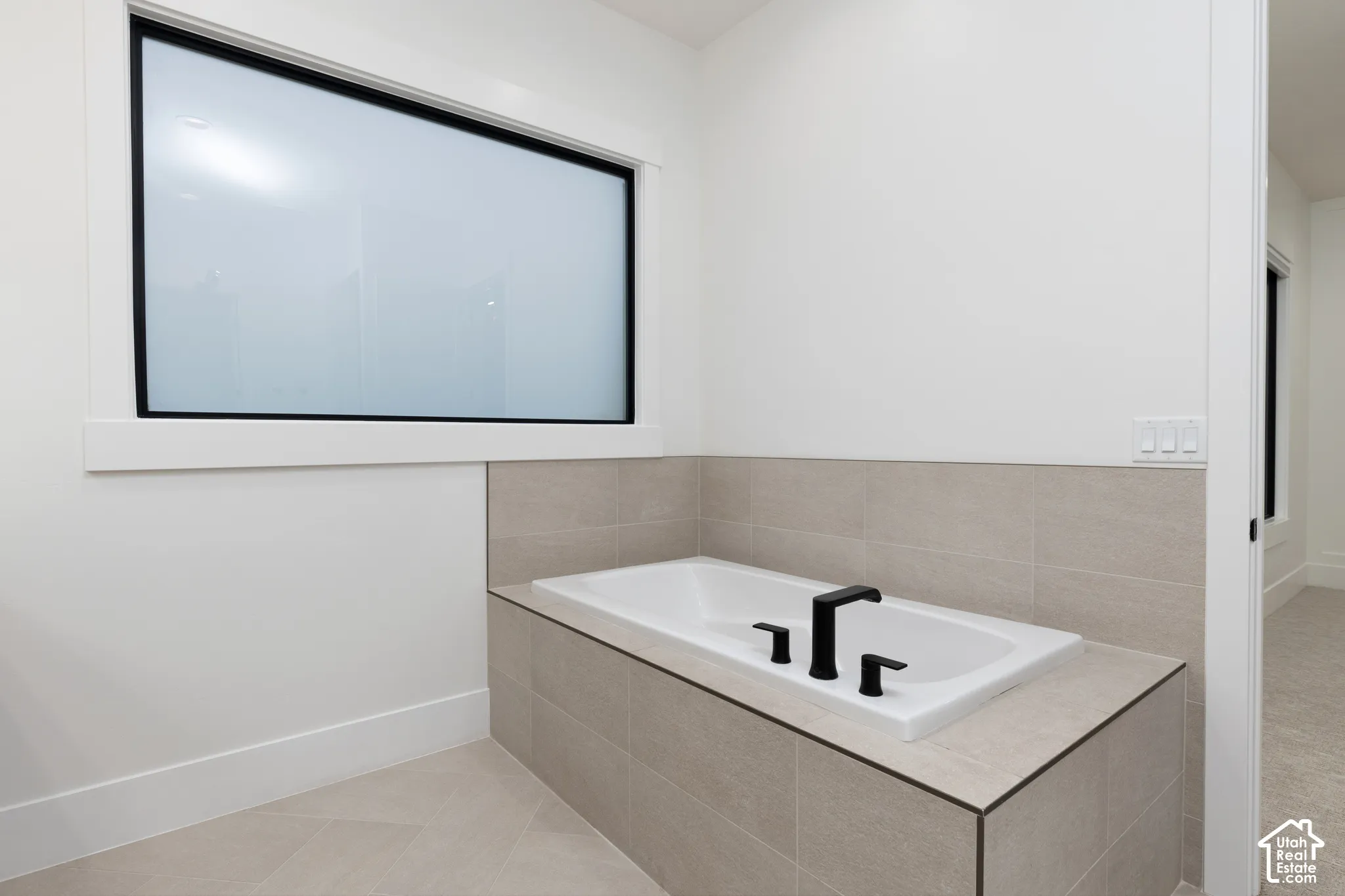 Built-in soaking tub with a matte black deck-mounted faucet, surrounded by neutral-toned ceramic tile and a frosted window with black trim