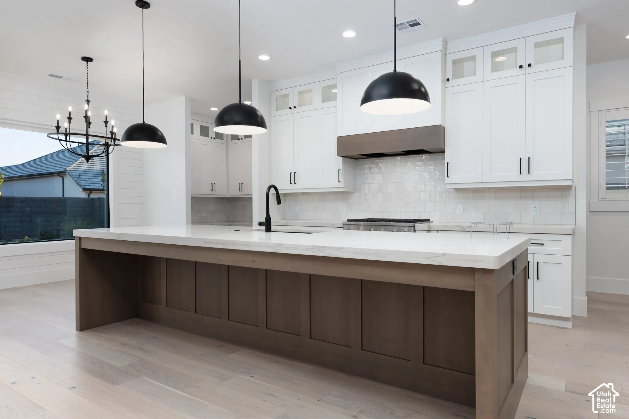 Kitchen featuring an expansive island with stone-surface countertop, white cabinetry with glass-front uppers, matte black plumbing fixtures, wood-finish flooring, and industrial-style pendant lighting