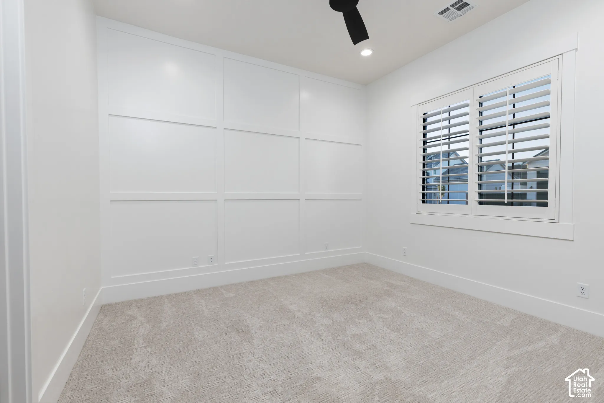 Room featuring white wall-paneling millwork, carpeting, plantation shutters, and a matte black ceiling fan