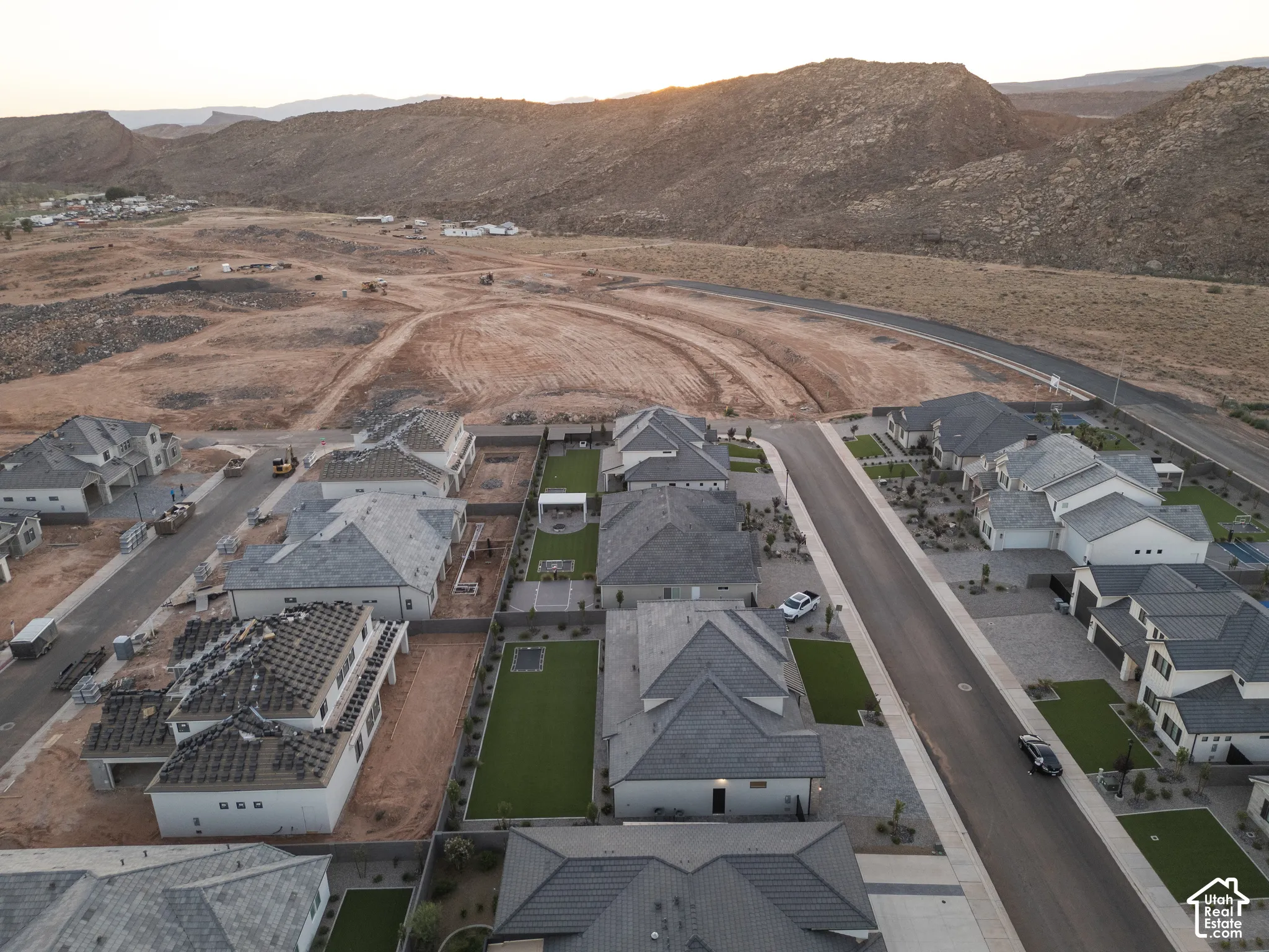 Aerial perspective of a residential development featuring single-story dwellings with pitched roofs, manicured artificial turf yards, paved walkways, and direct mountain backdrops