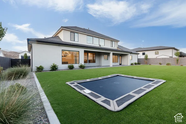 Stucco exterior featuring a covered patio, recessed in-ground trampoline, synthetic turf lawn, and perimeter masonry fencing