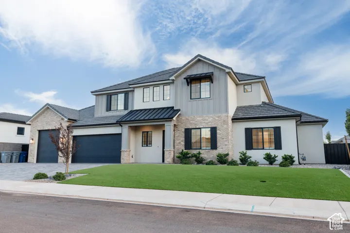 Two-story residence featuring a combination of light-toned stone veneer and smooth stucco exterior, dark metal roof accents, shutter-trimmed windows, and a multi-car garage with stone-paved driveway