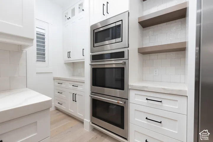 Custom white cabinetry with matte black hardware, stainless steel built-in double ovens and microwave, stone countertops, square-profile tile backsplash, and wood-finish flooring