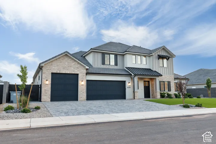 Multi-level exterior featuring a combination of brick and vertical siding, concrete paver driveway, three-car garage, and metal roof accents